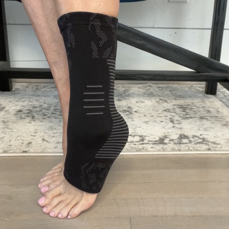 A black NanoSock worn on one foot and photographed from the side at floor level indoors, showing the patterned knit, the ribbed compression panels running up the ankle, the fully enclosed heel, and the bare toes coming out the front of the open toe box.